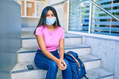 Tıp maskesi takan genç Latin öğrenci üniversitenin merdivenlerinde oturuyor..