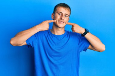 Günlük mavi tişört giyen, neşeli gülümseyen ve parmaklarıyla ve ağzıyla işaret eden genç beyaz bir adam. diş sağlığı kavramı. 