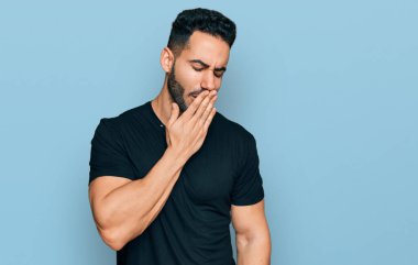 Hispanic man with beard wearing casual black t shirt bored yawning tired covering mouth with hand. restless and sleepiness. 