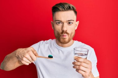 Young redhead man holding toothbrush with toothpaste and glass of water making fish face with mouth and squinting eyes, crazy and comical. 