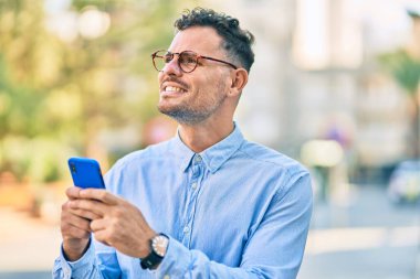 Genç İspanyol işadamı şehirde akıllı telefon kullanarak mutlu bir şekilde gülümsüyor..