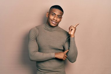 Balıkçı yaka kazak giyen, yüzünde kocaman bir gülümseme olan, eli ve parmağı kameraya bakan genç bir zenci.. 