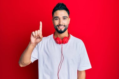 Young man with beard listening to music using headphones showing and pointing up with finger number one while smiling confident and happy. 