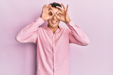 İspanyol asıllı genç adam sıradan kıyafetler giyiyor dürbün gibi jest yapıyor, gözleri parmaklarından bakıyor. çılgın ifade. 