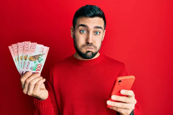 Homem Hispânico Jovem Usando Smartphone Segurando Notas Nova Zelândia Dólares — Fotografia de Stock