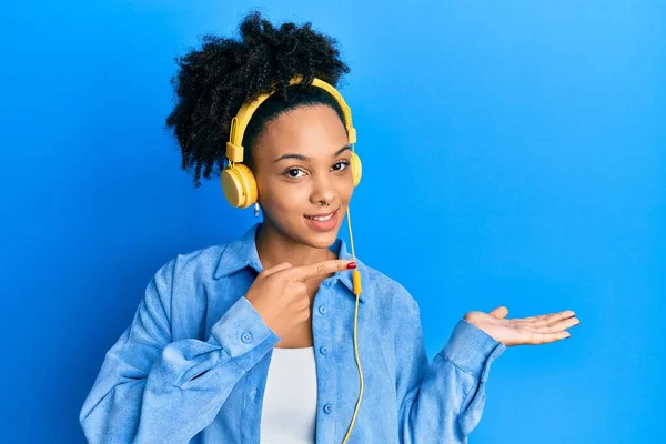 年轻的非洲裔美国女孩一边用耳机听音乐 一边用手拿着相机 用手指指指 一边惊讶地微笑着 — 图库照片