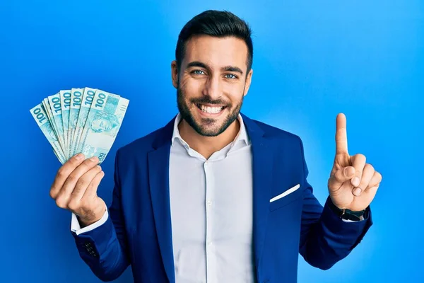 ブラジルの銀行券を持っているビジネススーツを身に着けている若いヒスパニック系のビジネスマンは幸せな顔でアイデアや質問指差し指で笑顔 ナンバーワン — ストック写真