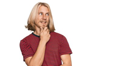 Sarışın, uzun saçlı, sıradan çizgili tişörtlü, elleri çenesinde soru soran, düşünceli bir beyaz adam. Düşünceli bir yüzle gülümsüyor. şüphe kavramı. 