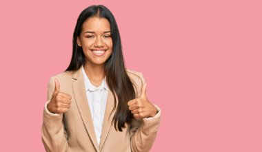 Güzel İspanyol kadın, iş ceketi giyiyor, başarı işareti yapıyor, elleriyle olumlu jest yapıyor, gülümsüyor ve mutlu. Neşeli ifade ve kazanan jest. 