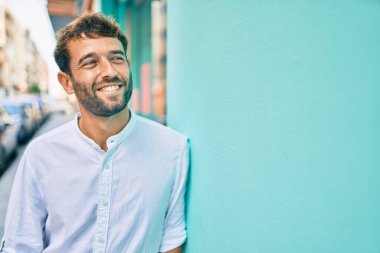 Güneşli bir günde günlük beyaz gömlek giyen sakallı yakışıklı adam dışarıda mutlu bir şekilde gülümsüyor.