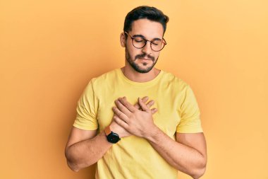 Gündelik kıyafetler ve gözlük takan genç İspanyol adam elleri göğsünde, gözleri kapalı ve yüzünde minnet dolu bir jest ile gülümsüyor. sağlık kavramı. 