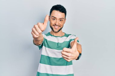 Latin asıllı genç adam gündelik giysiler giyip olumlu jestler yapıyor. Elleriyle gülümsüyor ve başarı için seviniyor. kazanan jest. 