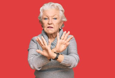 Kış süveteri giyen yaşlı gri saçlı kadın kollarını kavuşturup negatif işaret veriyor, kızgın yüz ifadesi takınıyor. 