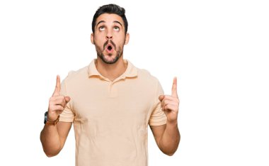 Gündelik giysiler giyen genç İspanyol adam şaşırmış ve şaşırmış bir şekilde yukarı bakıyor ve parmaklarıyla işaret ediyor ve kollarını kaldırıyor.. 