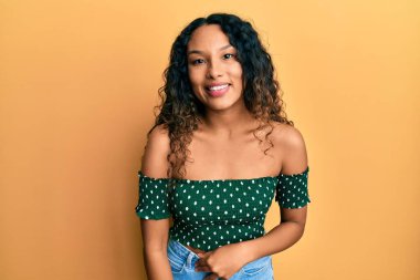 Gündelik kıyafetler giyen genç Latin kadın pozitif ve mutlu görünüyor. Duruyor ve gülümsüyor. Kendine güvenen bir gülümsemeyle dişlerini gösteriyor. 