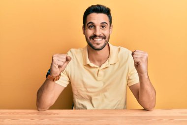 Latin asıllı genç adam, gündelik giysiler içinde masada oturmuş kollarını kaldırıp, gözleri açık bir şekilde başarıyı kutluyor ve şaşırıyor. Kazanan konsept. 
