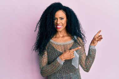 Orta yaşlı Afro-Amerikalı kadın günlük kıyafetler giyiyor gülümsüyor ve kameraya bakıyor iki eliyle ve parmaklarıyla yan tarafa bakıyor.. 