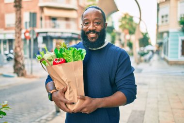 Sakallı Afrikalı Amerikalı bir adam elinde süpermarketten alınmış kağıt poşetler tutuyor.