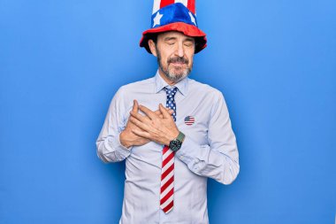 Orta yaşlı, yakışıklı, vatansever, şapkalı ve kravatlı, mavi arka planda elleri göğsünde, gözleri kapalı, yüzünde minnettarlıkla gülümseyen bir adam. Sağlık kavramı.