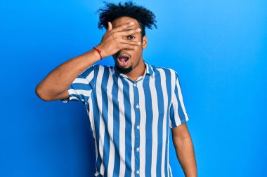 Sakallı, sıradan çizgili tişörtü olan, yüzünü ve gözlerini kapayarak utangaç ifadesiyle parmaklarından içeri bakan Afro-Amerikalı genç bir adam.. 