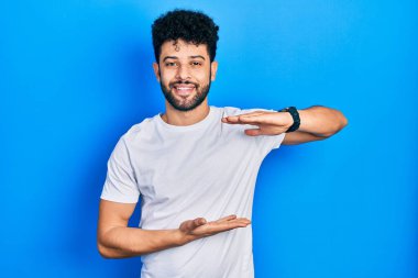 Sakallı, sıradan beyaz tişörtlü, elleri büyük ve büyük boy işareti gösteren genç bir Arap. Ölçü sembolü. Kameraya bakıp gülümsüyor. ölçüm kavramı. 