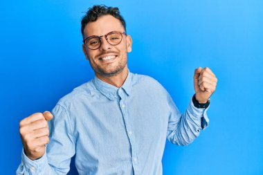 Gündelik kıyafetler giyen ve gözlük takan genç İspanyol adam mutlu ve neşeli dans ediyor, gülümsüyor rahat ve kendinden emin bir şekilde müzik dinliyor. 