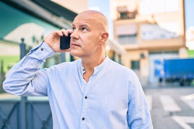 Middle age bald man with serious expression talking on the smartphone at the city.