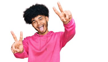 Afro saçlı, sıradan pembe kazaklı, ağzı açık gülümseyen ve parmaklarını iki eliyle zafer işareti yapan Afro-Amerikan genç adam. İki numara.. 