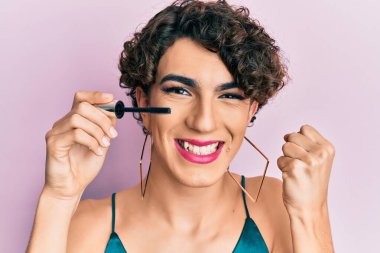Young man wearing woman make up holding eyelash mascara screaming proud, celebrating victory and success very excited with raised arm 