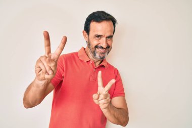 Orta yaşlı, sıradan kıyafetler giyen, gülümseyen, parmaklarını zafer işareti yaparken gösteren İspanyol bir adam. İki numara.. 