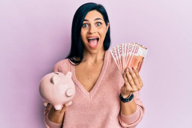 Young caucasian woman holding norwegian krona banknotes and piggy bank celebrating crazy and amazed for success with open eyes screaming excited. 