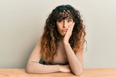 Latin kökenli genç bir kız, gündelik giysiler içinde masaya oturmuş, kollarını kavuşturarak depresyondan sıkılmış ve yorgun göründüğünü düşünüyor.. 
