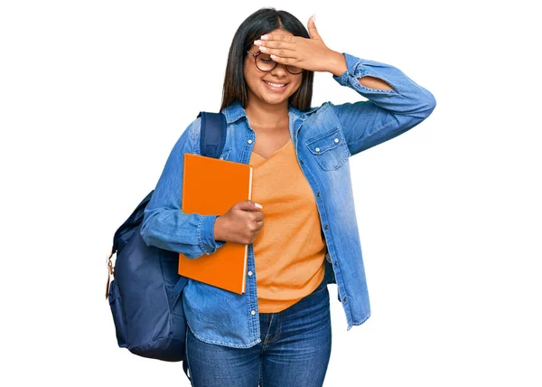 若いラテン語の少女学生のバックパックを着て 笑顔と驚きのために目をカバー顔に手で笑って本を保持します 視覚障害の概念 — ストック写真