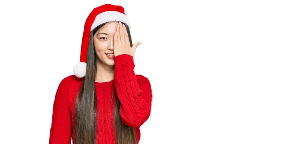 年轻的中国女人戴着圣诞帽 一只手捂住一只眼睛 脸上挂着自信的微笑 还有令人惊讶的情绪 — 图库照片