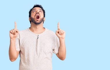 Gündelik giysiler giyen genç İspanyol adam şaşırmış ve şaşırmış bir şekilde yukarı bakıyor ve parmaklarıyla işaret ediyor ve kollarını kaldırıyor.. 
