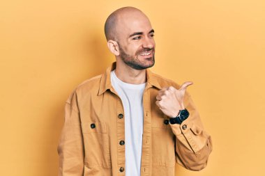 Genç kel bir adam, günlük kıyafetler giymiş, gülen yüzlü, gülümseyen ve baş parmağıyla yan tarafı gösteren.. 