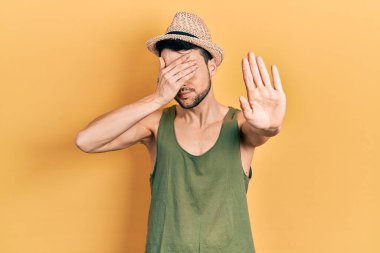 Yaz şapkası takan genç İspanyol adam elleriyle gözlerini kapatıyor ve üzgün ve korku ifadeleriyle jest yapıyor. utangaç ve olumsuz kavram. 