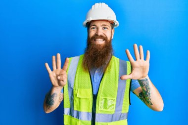 Uzun sakallı, güvenlik kaskı takan ve yansıtıcı ceketli kızıl saçlı adam dokuz numarayı gösterip işaret ederken kendinden emin ve mutlu bir şekilde gülümsüyor.. 