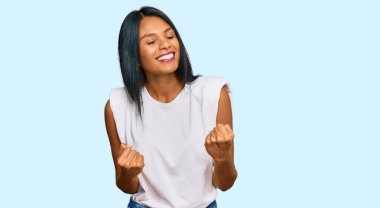 Genç Afro-Amerikalı kadın gündelik giysiler giyip çok mutlu ve heyecanlı bir şekilde kollarını kaldırıp, gülümseyerek ve başarı için bağırarak kazanan bir jest yapıyor. kutlama konsepti. 