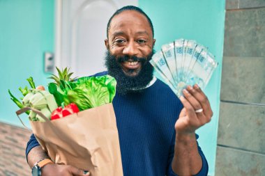 Elinde süpermarketten alınmış kağıt poşetlerle sakallı Afrikalı Amerikalı bir adam 100 Brezilya banknotunu gösteriyor.
