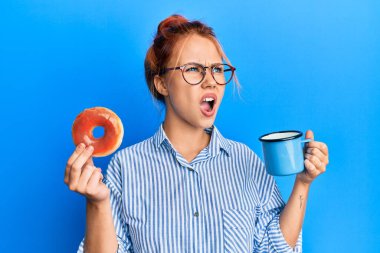 Genç kızıl saçlı kadın elinde çikolatalı çörek ve kahveyle kahvaltı yapıyor kızgın ve öfkeli çığlıklar atıyor, öfkeyle bağırıyor. öfke ve agresif konsept. 