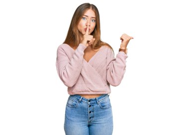 Beautiful blonde woman wearing casual winter pink sweater asking to be quiet with finger on lips pointing with hand to the side. silence and secret concept. 