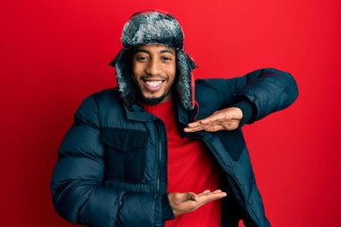 Sakallı, kışlık şapkalı ve paltolu, elleri büyük ve büyük bir işaret gösteren Afrikalı genç bir Amerikalı. Ölçü sembolü. Kameraya bakıp gülümsüyor. ölçüm kavramı. 