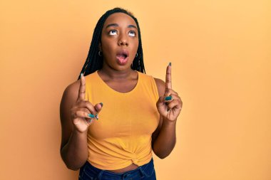 Genç Afrikalı Amerikalı kadın gündelik kıyafetler giyip şaşkın şaşkın yukarı bakıyor ve parmaklarıyla işaret ediyor ve kollarını kaldırıyor.. 