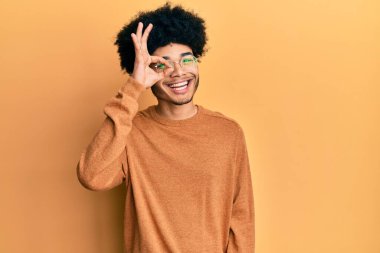 Afro saçlı, sıradan bir kazak giyen, el hareketleriyle gülümseyen, mutlu yüzüyle parmaklarının arasından bakan Afro Amerikalı genç bir adam.. 