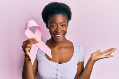 Genç Afro-Amerikalı kız elinde pembe kanser kurdelesi, zaferi mutlu bir gülümsemeyle kutluyor ve kazanan el kaldırarak ifade ediyor. 