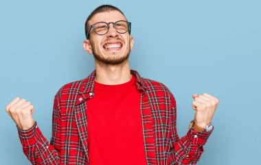 İspanyol genç adam gündelik kıyafetler giyip çok mutlu ve heyecanlı bir şekilde kollarını kaldırıp, gülümseyerek ve başarı için bağırarak kazanan jestler yapıyor. kutlama konsepti. 