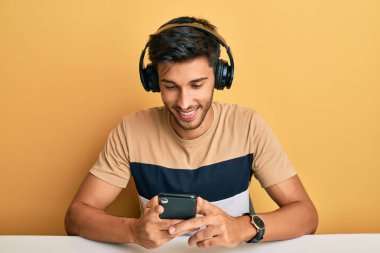 Akıllı telefon kullanan, kulaklık takan, pozitif ve mutlu görünen, kendine güvenen, gülümseyen, dişlerini gösteren genç yakışıklı bir adam. 