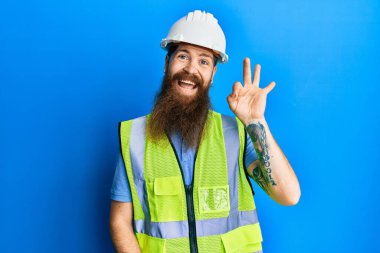 Uzun sakallı, güvenlik kaskı takan ve yansıtıcı ceketli kızıl saçlı adam gülümseyerek el ve parmaklarıyla 