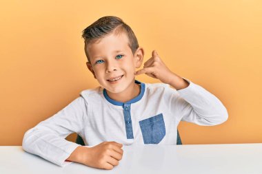 Sevimli beyaz çocuk, günlük giysiler içinde masaya oturmuş gülümsüyor telefonla el ve parmaklarıyla telefonla konuşuyor gibi yapıyor. iletişim kavramları. 